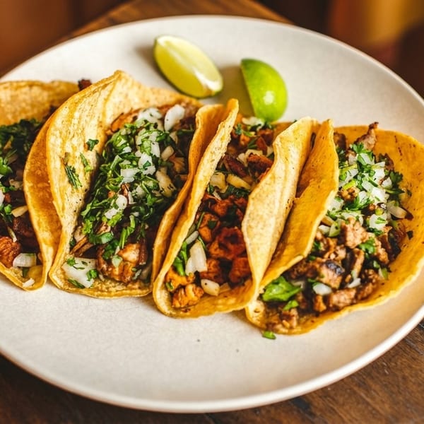 Five street tacos with meat, cilantro, and onions, served with lime wedges on a white plate.