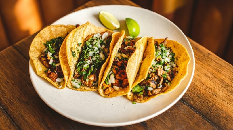 Five street tacos with meat, cilantro, and onions, served with lime wedges on a white plate.