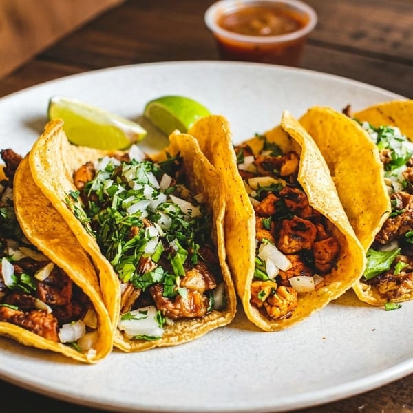 Four street tacos with meat, cilantro, and onions, served with lime wedges and salsa.