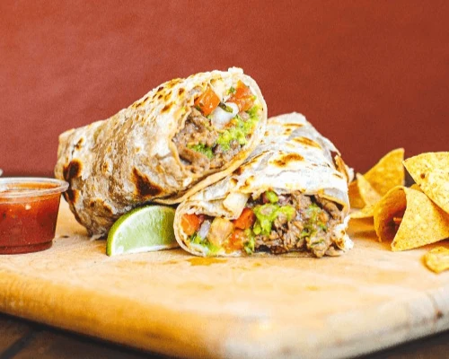 A sliced burrito filled with beef, guacamole, and salsa on a wooden board, accompanied by tortilla chips, a lime wedge, and a small cup of salsa.