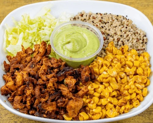 A white bowl contains grilled chicken, corn, quinoa, shredded lettuce, and creamy green sauce in a cup. The dish looks vibrant and fresh.