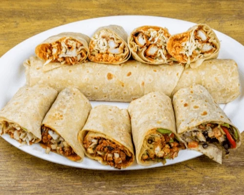 A white plate with assorted wraps cut in halves and rolled. They contain fillings like chicken, cheese, veggies, placed on a rustic wooden table, conveying a fresh and flavorful meal.
