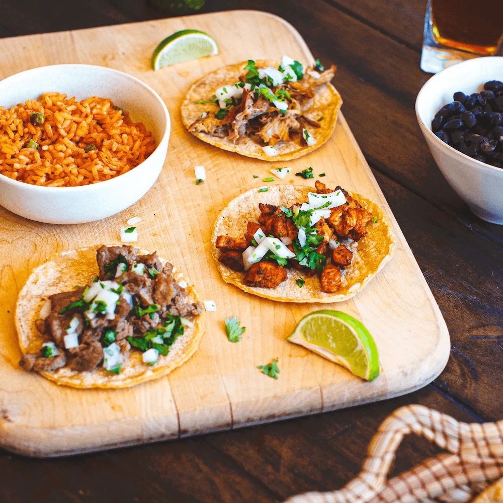 Three tacos with varied meats and fresh cilantro sit on a wooden board, accompanied by lime wedges, a bowl of orange rice, and black beans on a rustic table.