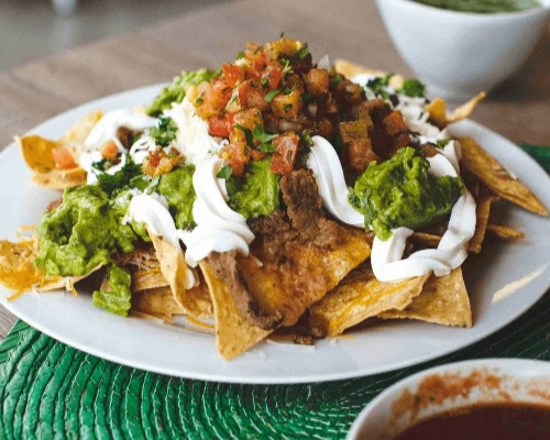 A plate of nachos topped with guacamole, salsa, sour cream, and seasoned beef, set on a textured green mat. A bowl of dip is blurred in the background.