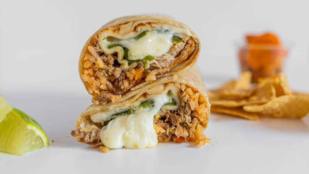 Two halves of a stuffed burrito with melted cheese, rice, and meat are stacked, with a lime wedge and tortilla chips blurred in the background.
