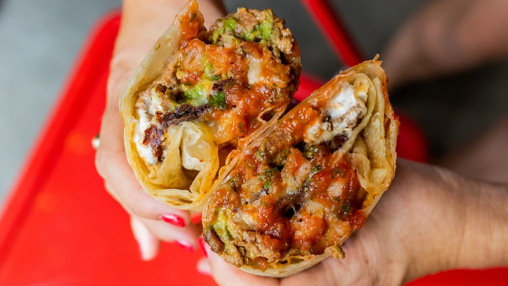 Close-up of a cheezy, vibrant burrito cut in half, revealing layers of meat, guacamole, melted cheese, and red salsa, held by hands with red nail polish.