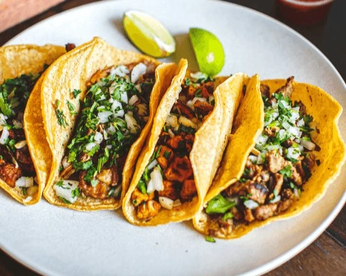 Four tacos with seasoned meat, chopped onions, and cilantro are arranged on a white plate. Two lime wedges are in the background, adding vibrancy.