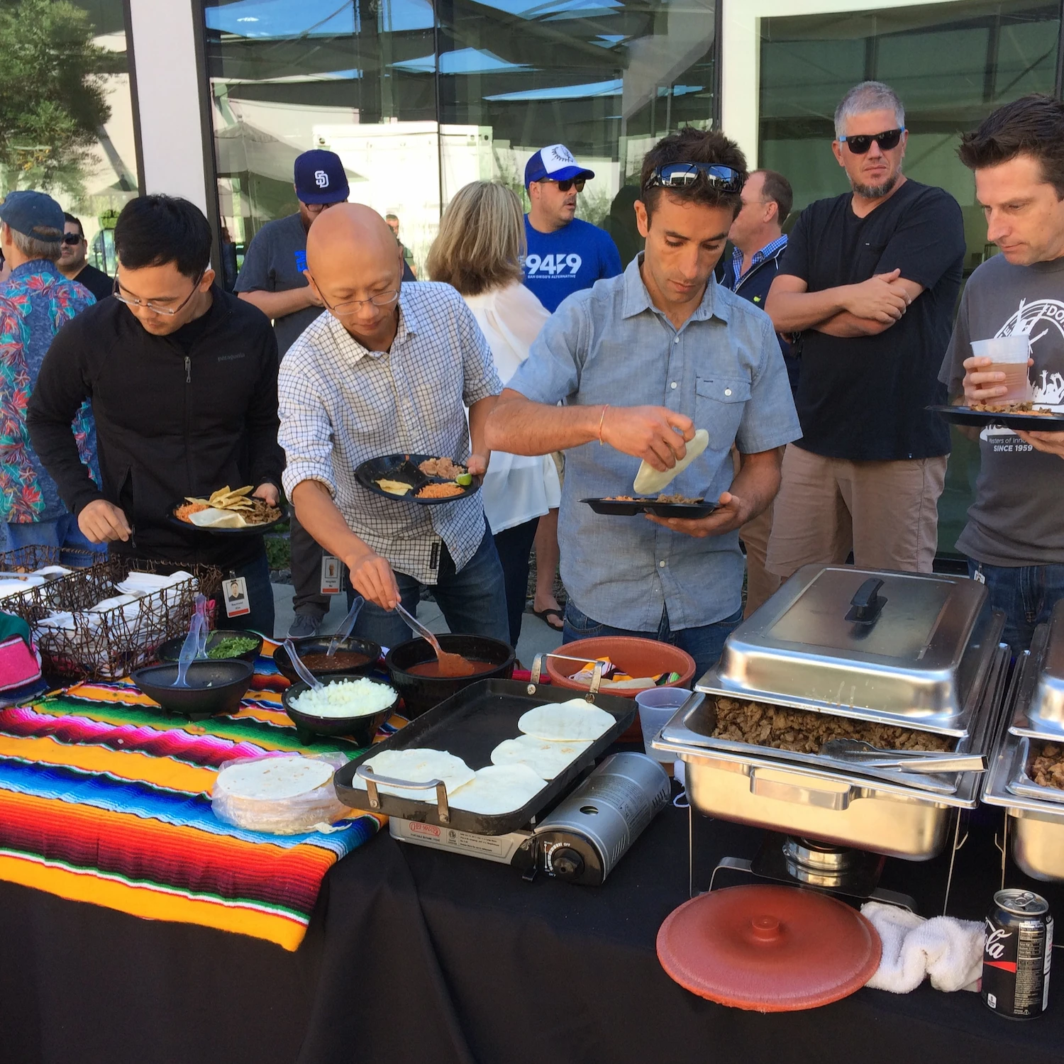 A group of people at an outdoor event serves themselves food from a buffet. A colorful blanket decorates the table, evoking a festive, relaxed atmosphere.