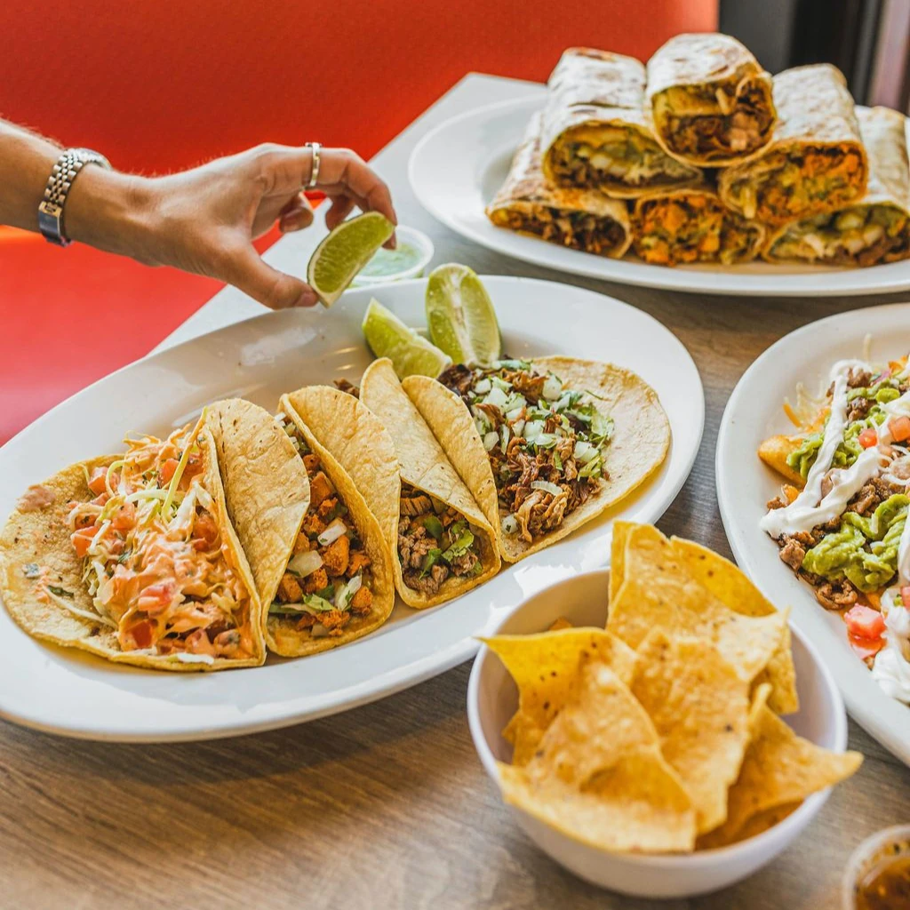 A platter of three assorted tacos with meat and fresh toppings garnished with lime wedges. A hand reaches for a lime. Surrounding dishes include wraps and a bowl of chips, creating a vibrant, inviting Mexican meal scene.