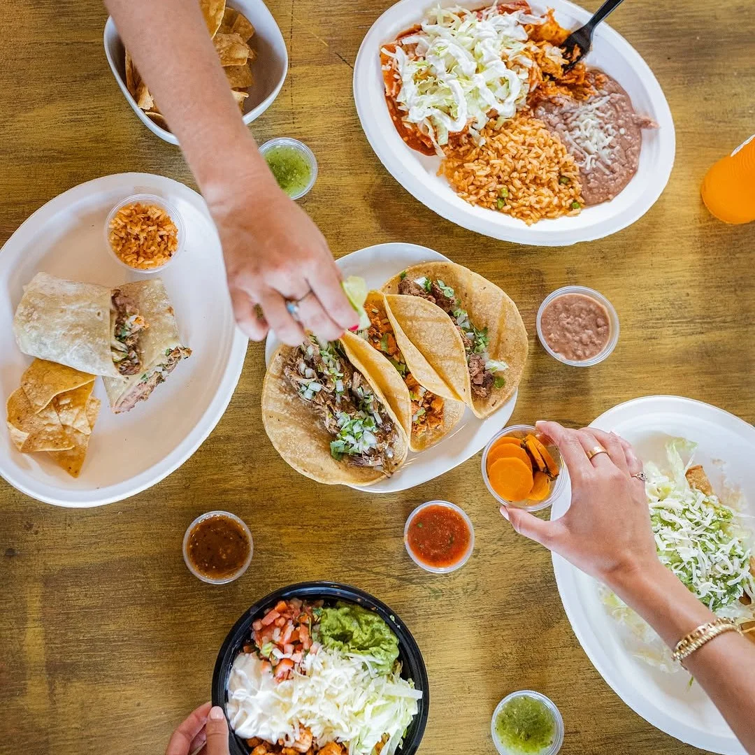 Aerial view of a table with Mexican dishes including tacos, burritos, and chips. Two hands reach for salsa and a drink, creating a lively, festive vibe.