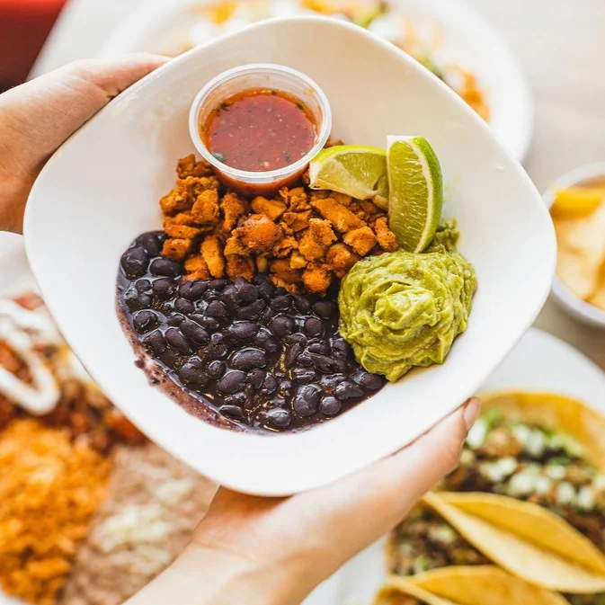 A colorful dish in a white bowl contains black beans, crumbled chorizo, creamy guacamole, lime wedges, and a small cup of salsa. Hands hold the bowl above a table.