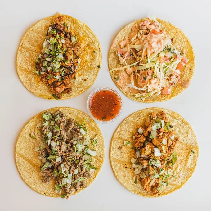 Four tacos on corn tortillas are arranged in a square around a small cup of red salsa. Each taco has different meat toppings with cilantro and onions.