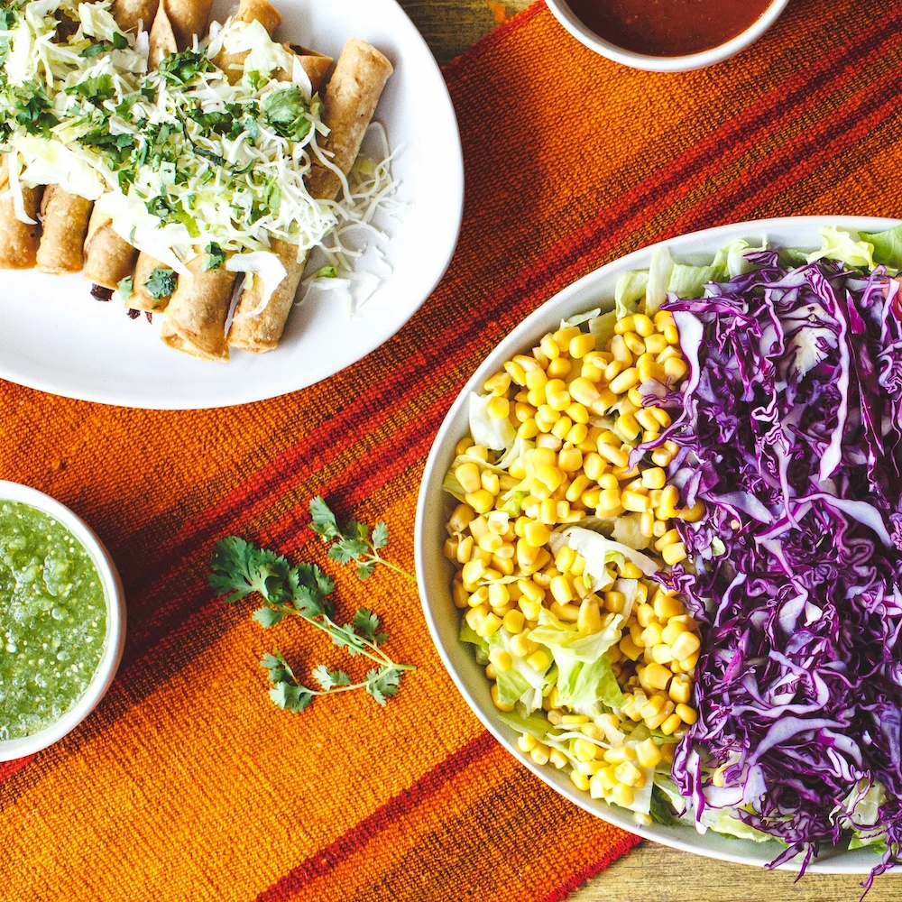 A vibrant Mexican meal on an orange striped mat. Crispy taquitos topped with cheese and cilantro, alongside a colorful salad with corn and purple cabbage.