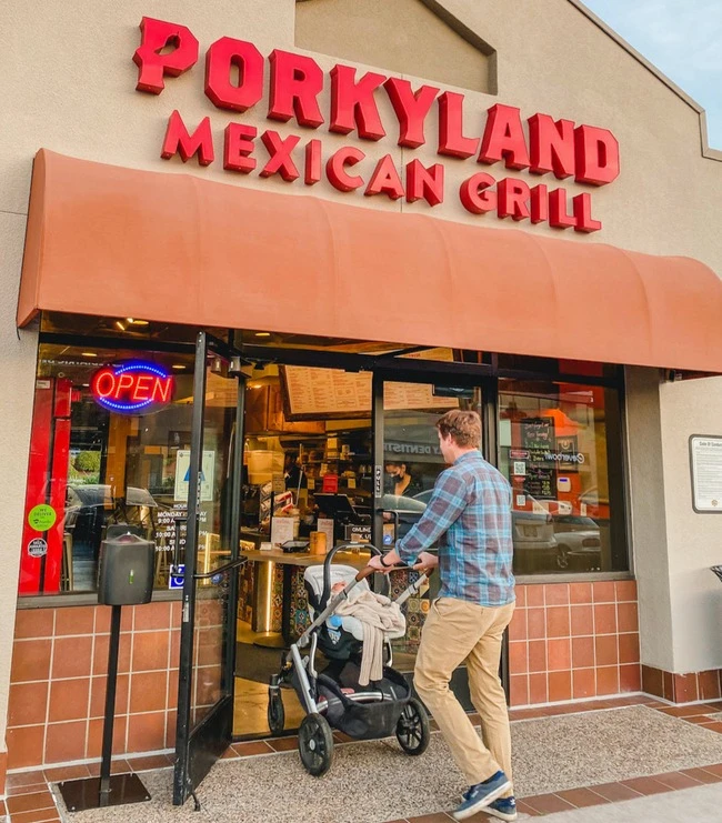 A man in a flannel shirt pushes a stroller into Porkyland Mexican Grill. The storefront features a red sign and neon "Open" sign, creating a welcoming vibe.