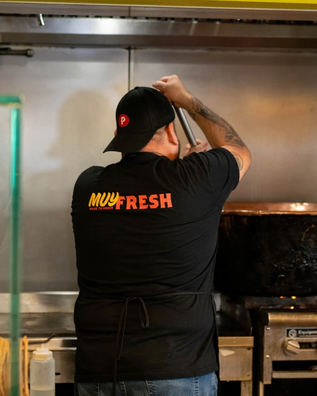 A cook in a black "Muy Fresh" shirt and cap works intently at a kitchen stove, stirring a large pot. The atmosphere is diligent and focused.