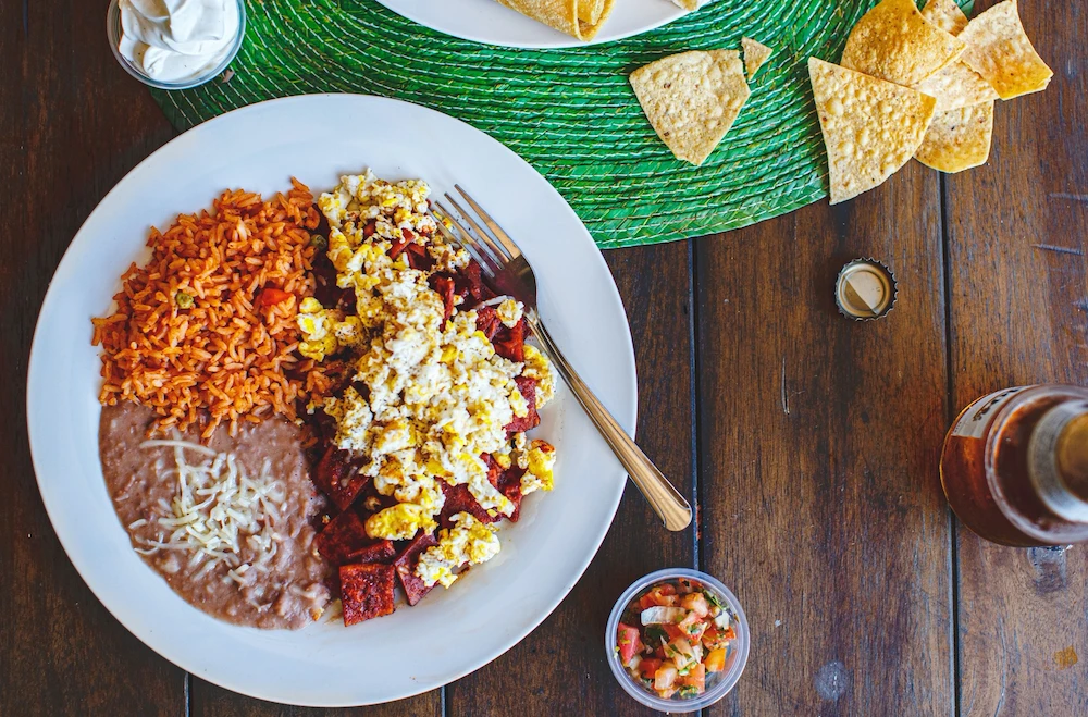 A vibrant Mexican meal on a wooden table: scrambled eggs with chorizo, refried beans, rice on a plate, tortilla chips, salsa, and hot sauce nearby.