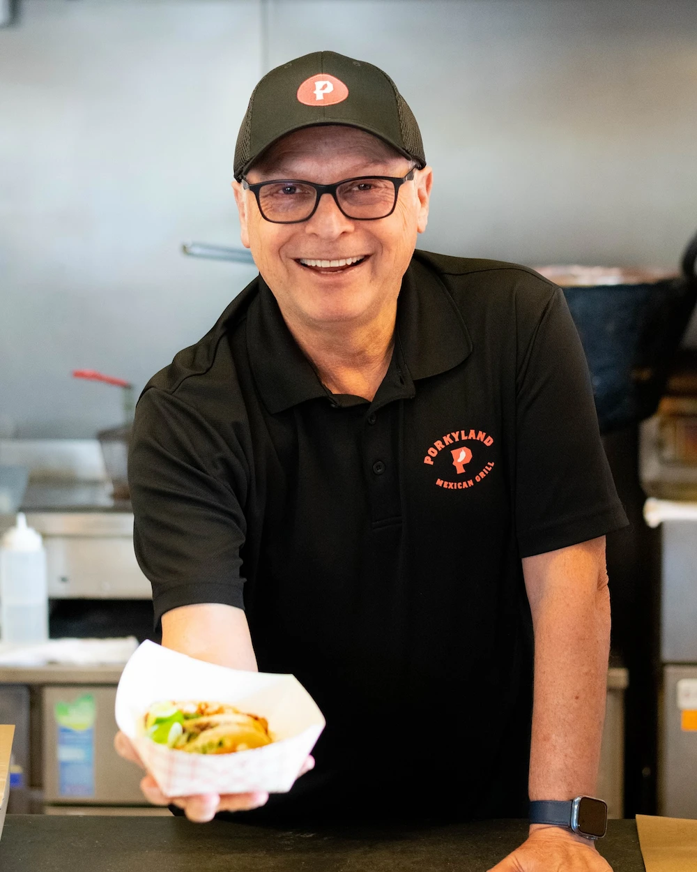 Smiling man in a black "Porkyland Mexican Grill" uniform and cap presents a taco in a white paper tray, conveying warmth and hospitality in a kitchen setting.