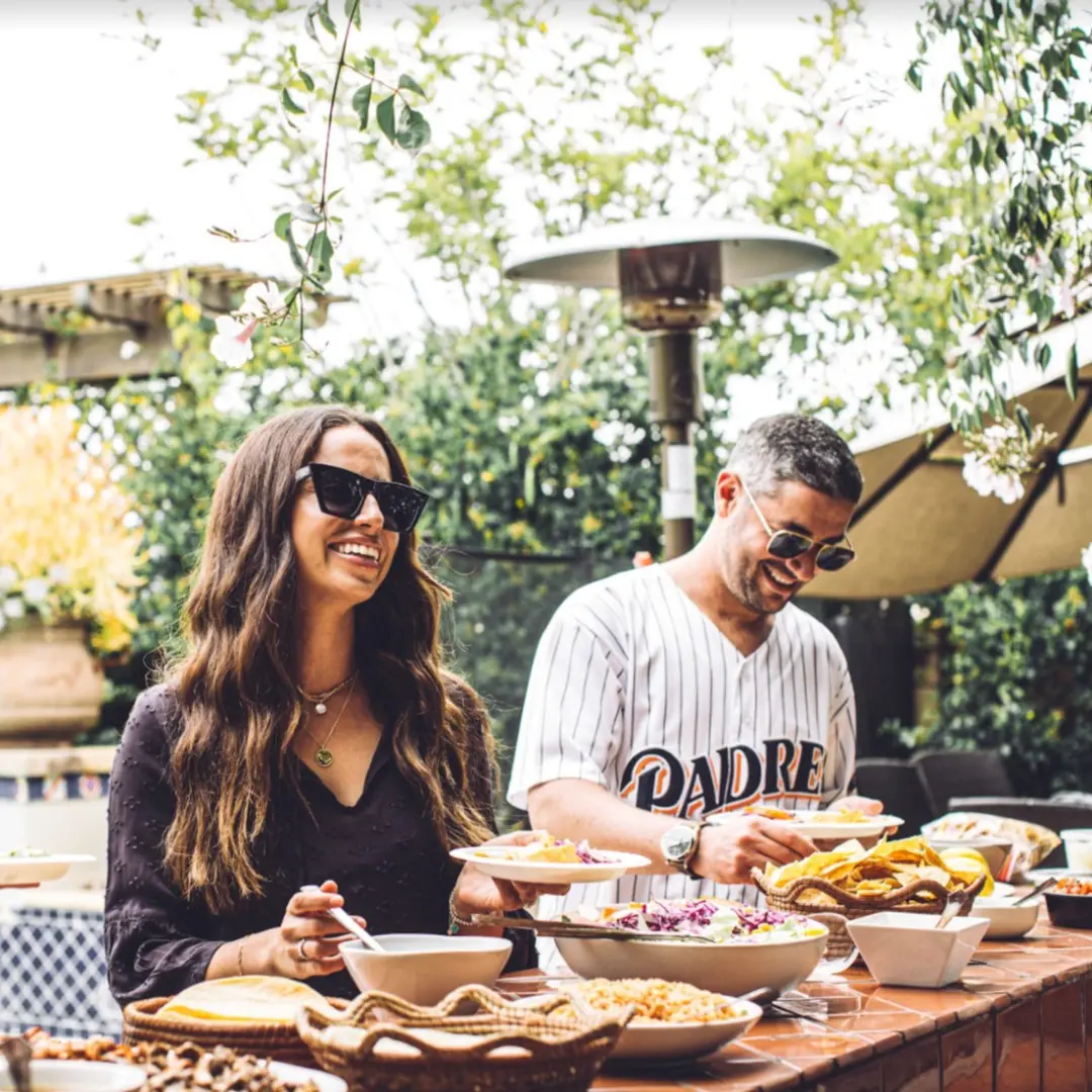 Two people enjoy an outdoor buffet, smiling and holding plates of food. The setting is bright and leafy, conveying a joyful, casual atmosphere.